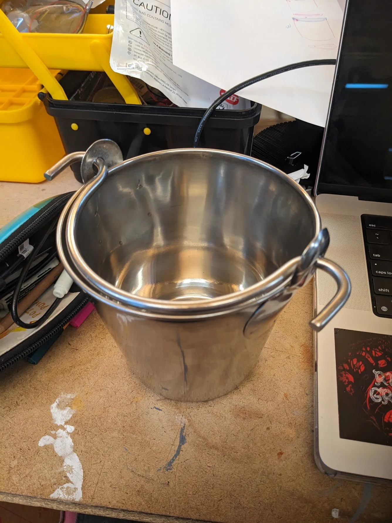 The replacement stainless steel pail with its wire handle, sitting on the workbench next to tools and a laptop.