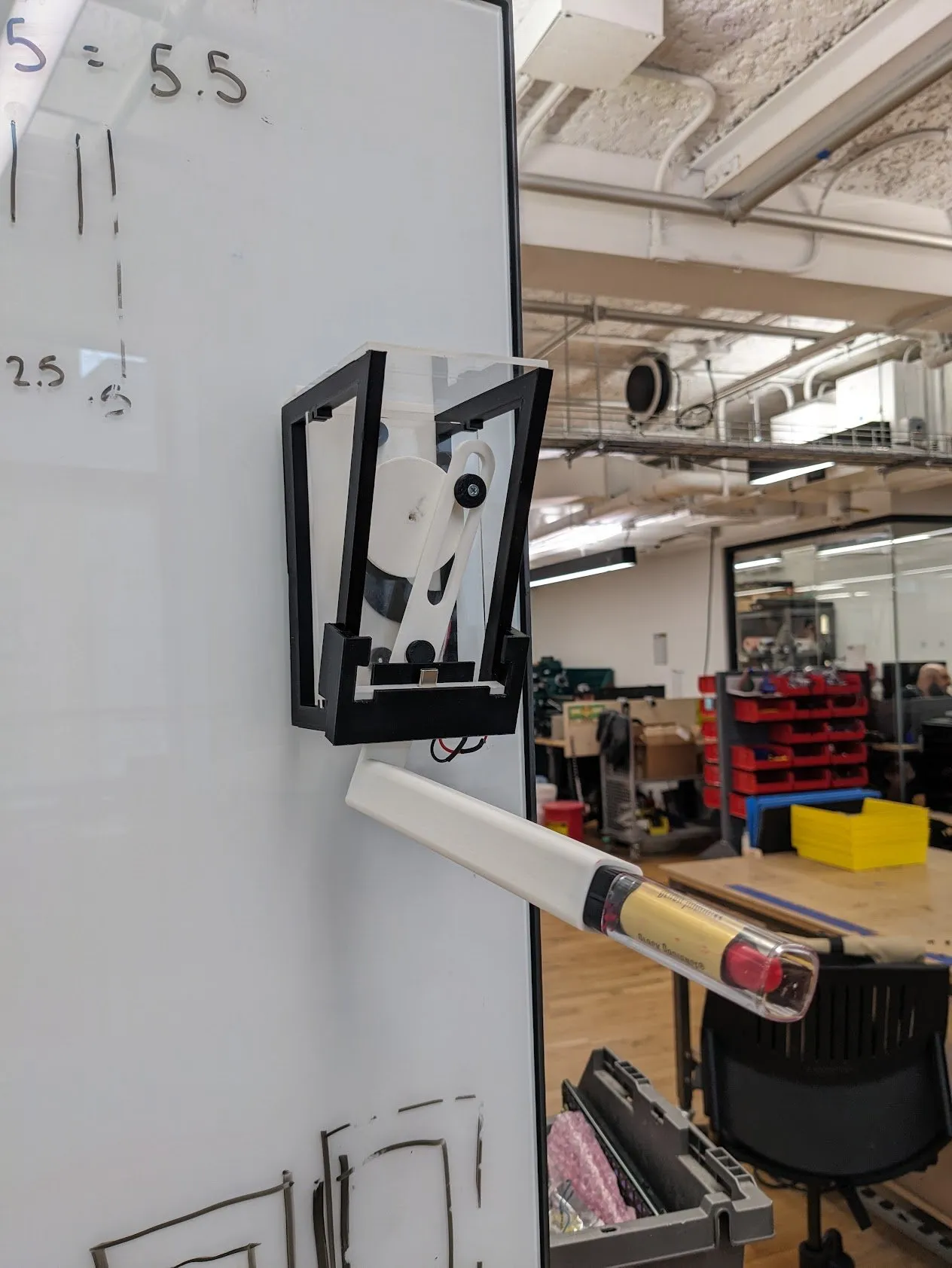 The finished frame suction-cupped to a glass partition with the phone clamped in and the lipstick arm extended, red test marks on the glass and "S = 5.5" written above.