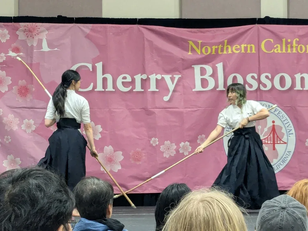 Two practitioners in keikogi and hakama demonstrating on stage at the Northern California Cherry Blossom Festival, naginata extended and crossing between them, an audience watching from below.