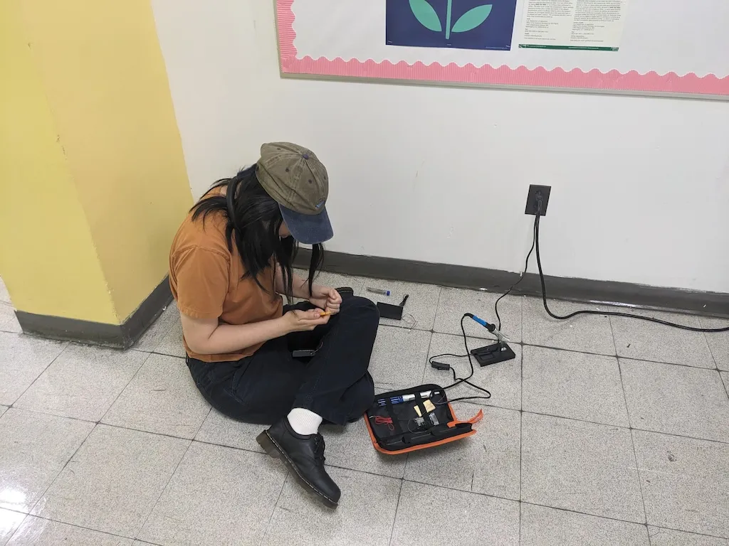 A photograph of an Asian woman sitting on the floor in the hallway of school soldering some pants.