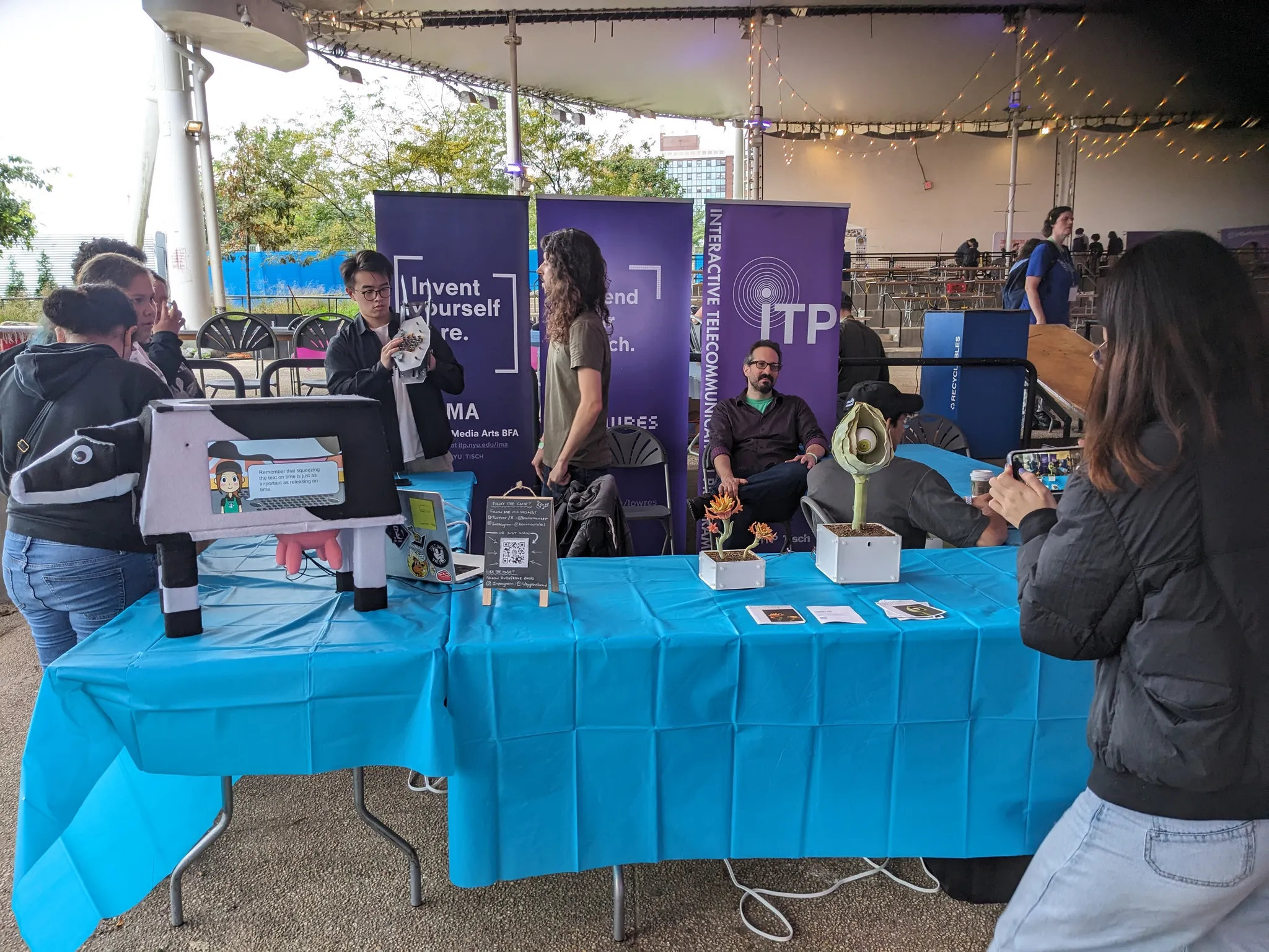 A photograph of the NYU ITP table at Coney Island Maker Faire, complete with violet banners and a cow and electronic plants.