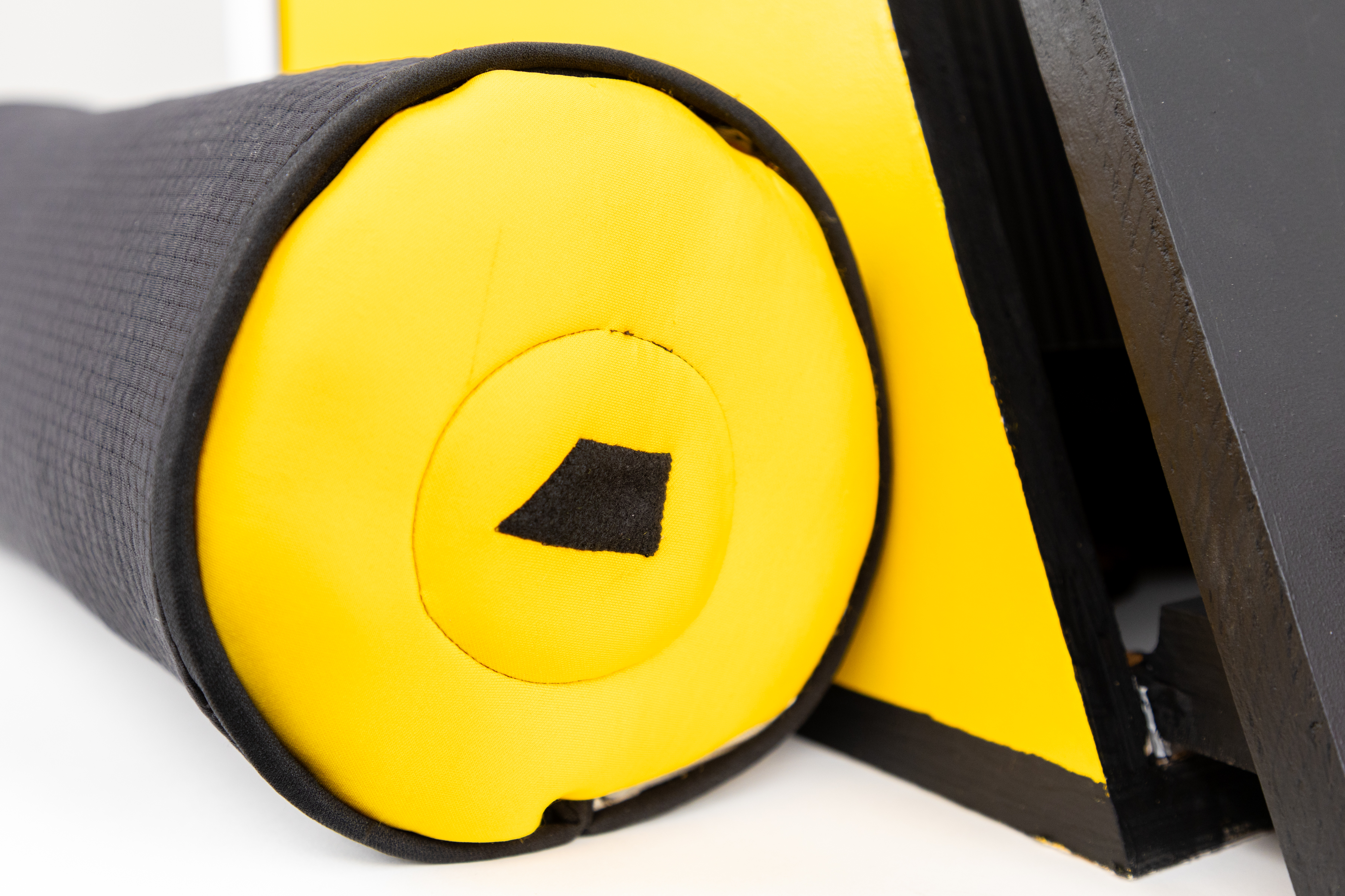 The image shows a close-up of a cylindrical object, likely a foam roller or similar fitness equipment. The object has a black textured surface on one side and a bright yellow end with a small black shape in the center. The yellow end is smooth and rounded, contrasting with the black material. In the background, there is a portion of a black structure, possibly part of a larger piece of equipment. The overall composition emphasizes the vibrant colors and textures of the roller.