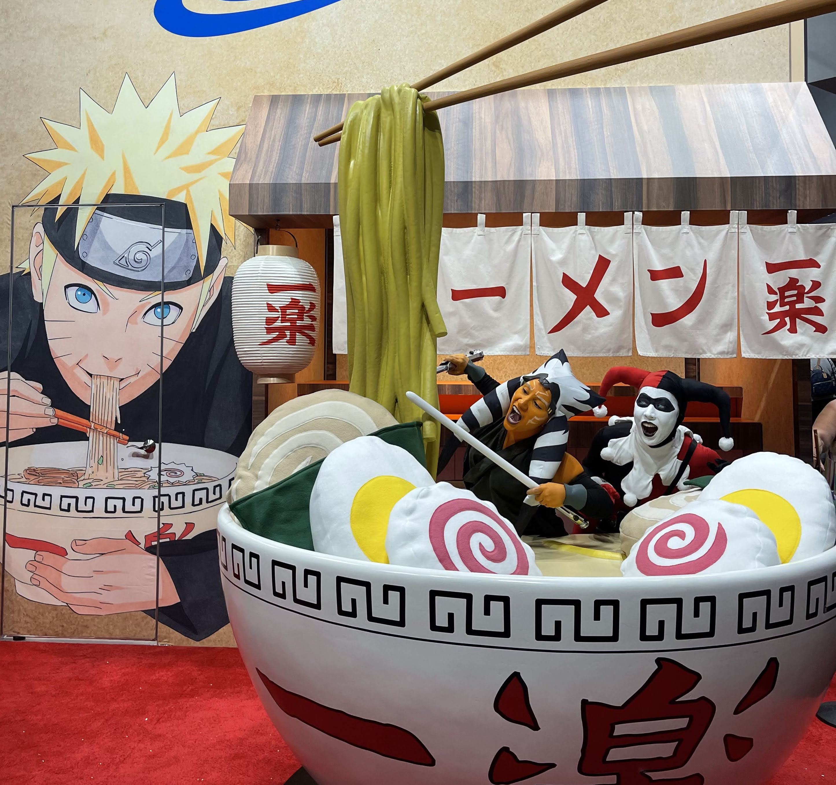 Photograph of a woman in a red and black harlequin outfit eating a giant fake bowl of ramen