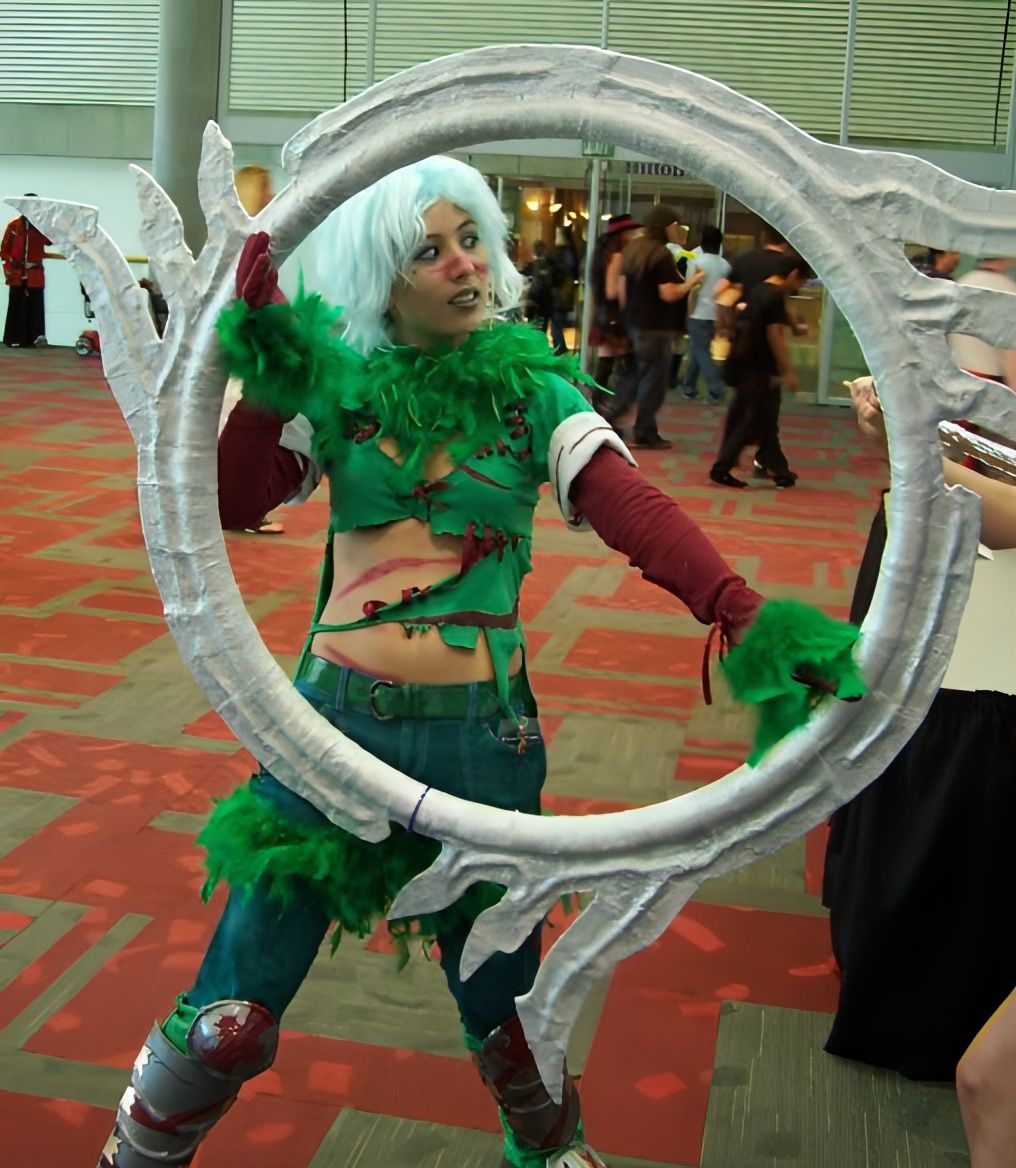 Photograph of a girl in a green feather outfit and blue hair holding a giant ring with blades