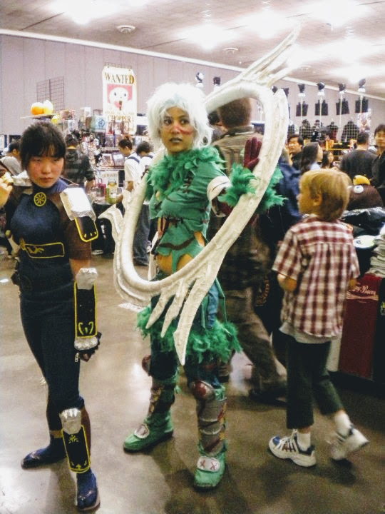 Photograph of a girl in a green feather outfit and blue hair holding a giant ring with blades standing next to a ninja