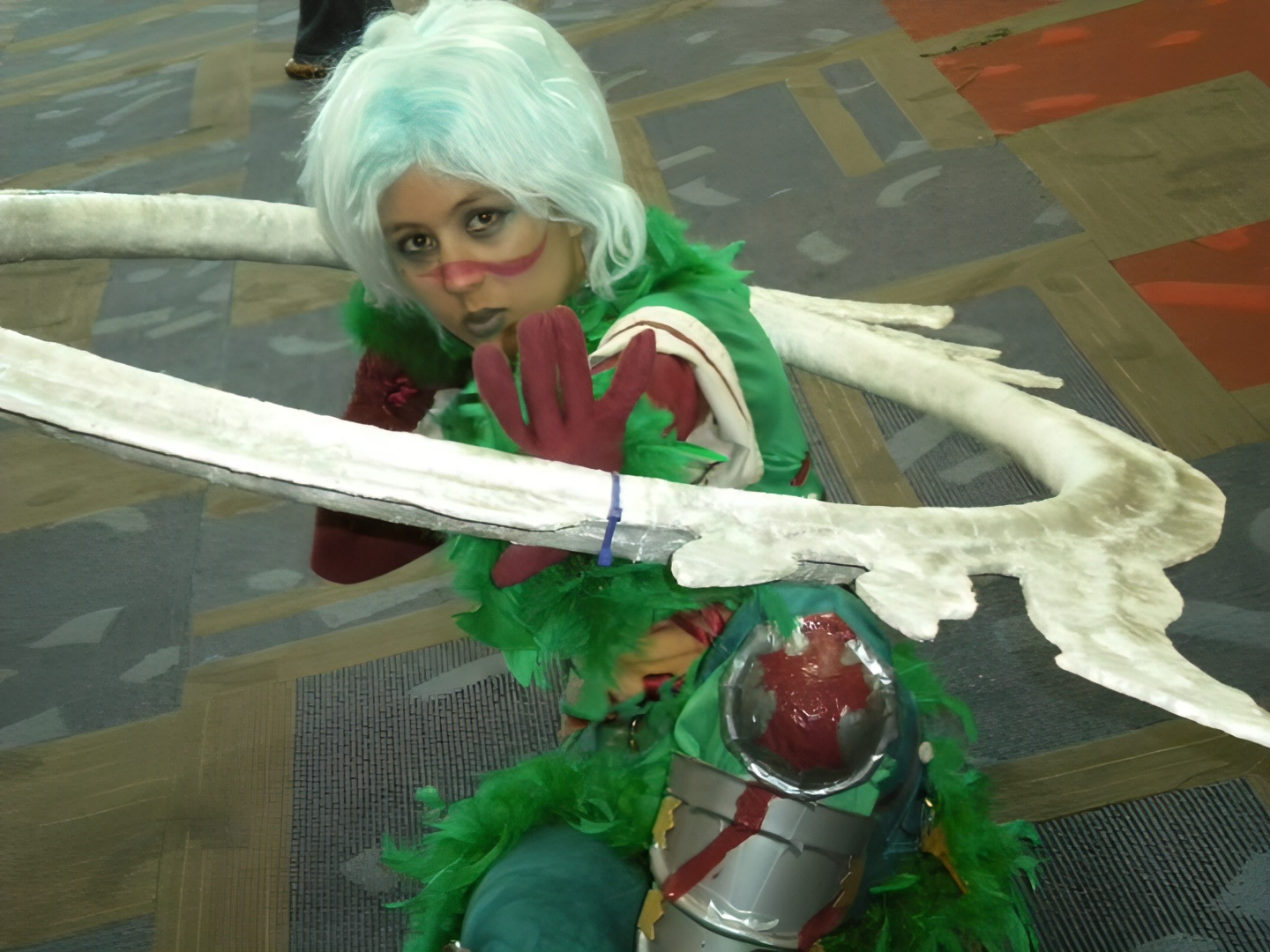 Photograph of a girl in a green feather outfit and blue hair pushing a giant ring with blades toward the camera